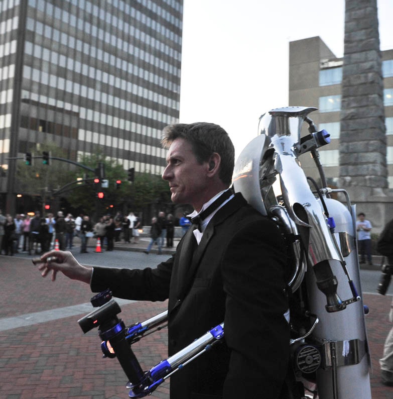 The Rocketman in a tuxedo with the rocketbelt strapped on, cigar in hand, city street behind him