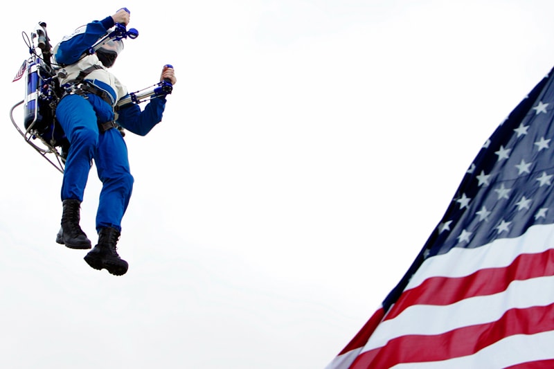 The Rocketman flying in a blue flight suit alongside a giant American flag