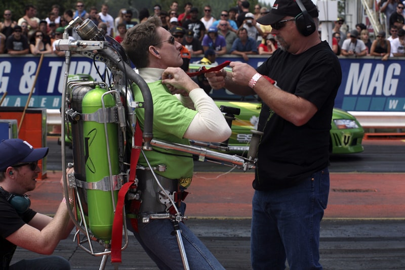 The Rocketman being strapped into the green rocketbelt at a drag racing event